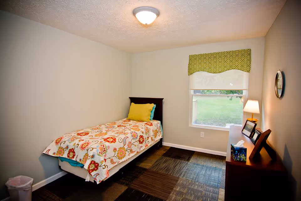 A small bedroom with a single bed covered in a floral patterned quilt and a yellow pillow. There is a window with a green patterned valance letting in natural light. A wooden dresser with a lamp, picture frames, and a tissue box is placed against the wall. The floor has a patchwork carpet design, and a small trash bin is in the corner.