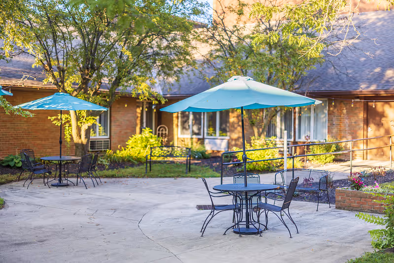 Outdoor patio area with round metal tables and chairs under blue umbrellas, surrounded by trees and greenery, with a brick building in the background.