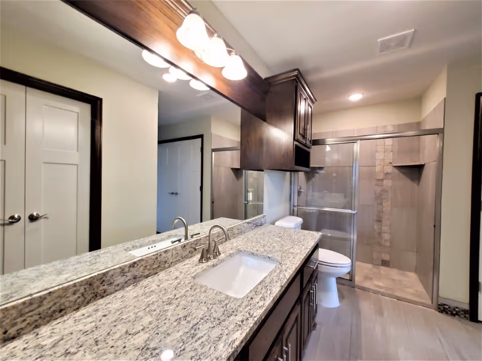 Well-lit bathroom with a granite sink vanity, large mirror, toilet, and glass-enclosed shower.