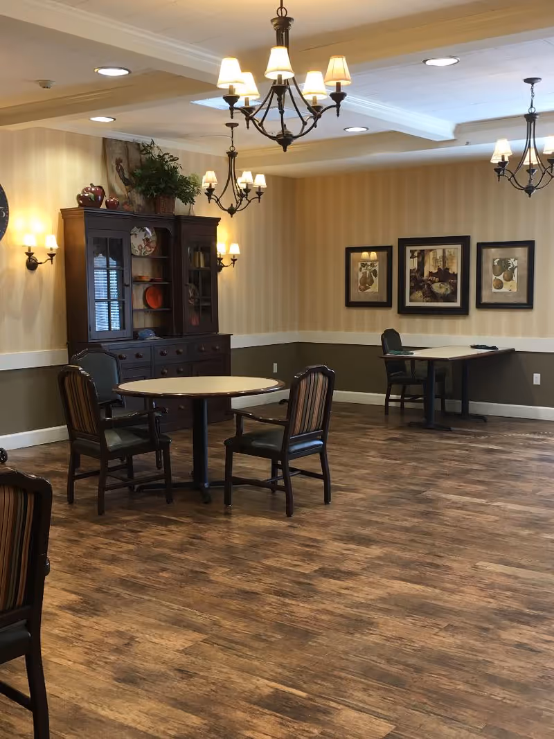 A warmly lit communal dining/activity room with round tables, chairs, chandeliers, a wooden cabinet, and framed wall art.
