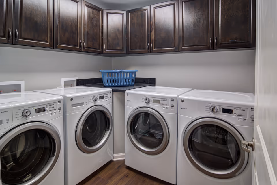 Laundry room with four front-loading washing machines and dryers lined up against the walls. Above the machines are dark wooden cabinets for storage, and a blue laundry basket is placed on the countertop in the corner. The room has wooden flooring and light gray walls.