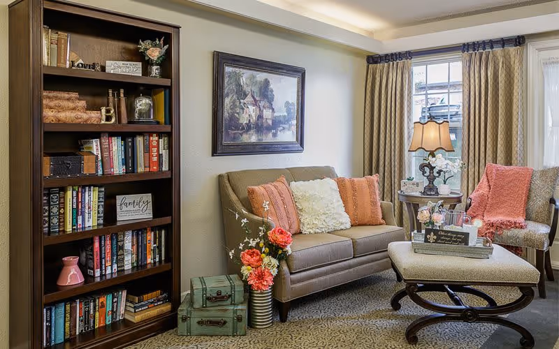 Cozy living room area with a beige sofa adorned with coral and white pillows, a cushioned ottoman coffee table, a side table with a lamp and flowers, a patterned armchair with a coral throw, a tall wooden bookshelf filled with books and decorative items, and a framed painting on the wall. The room has large windows with beige curtains letting in natural light.