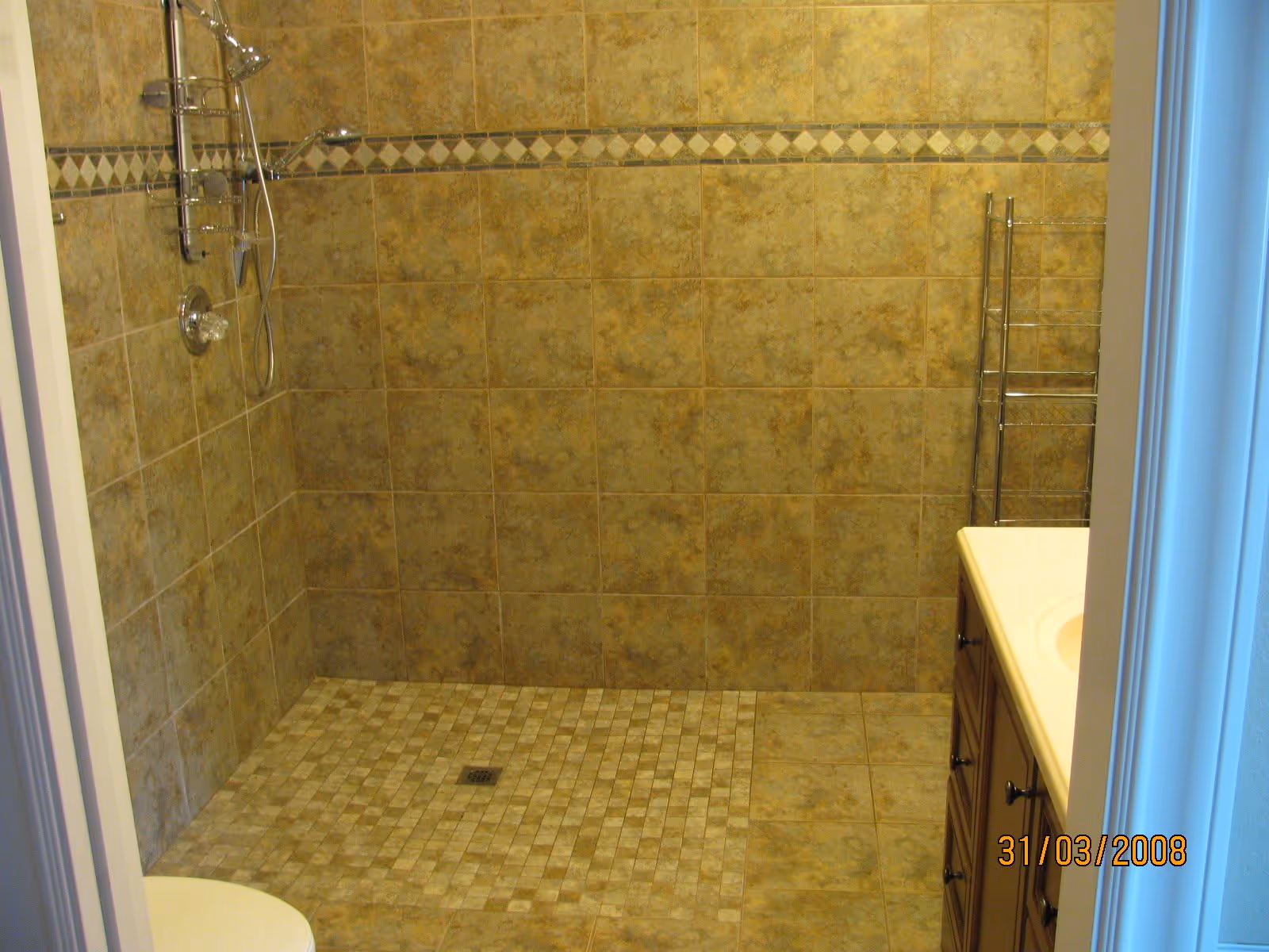 A tiled bathroom shower area with beige and brown tiles on the walls and floor. The shower has a chrome showerhead and a handheld shower wand. There is a small metal rack for storage and a wooden vanity with a white countertop visible on the right side.