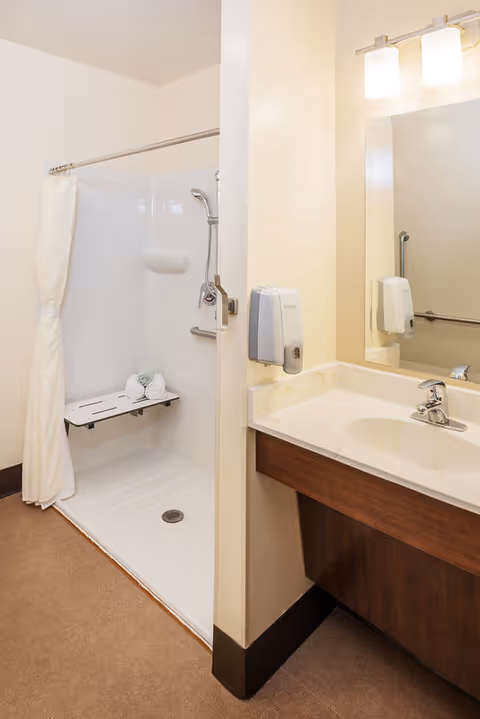A clean and accessible bathroom featuring a walk-in shower with a fold-down seat and a handheld showerhead. The shower has a white curtain and three rolled towels placed on the seat. Adjacent to the shower is a vanity with a sink, a large mirror, a soap dispenser mounted on the wall, and two overhead lights.
