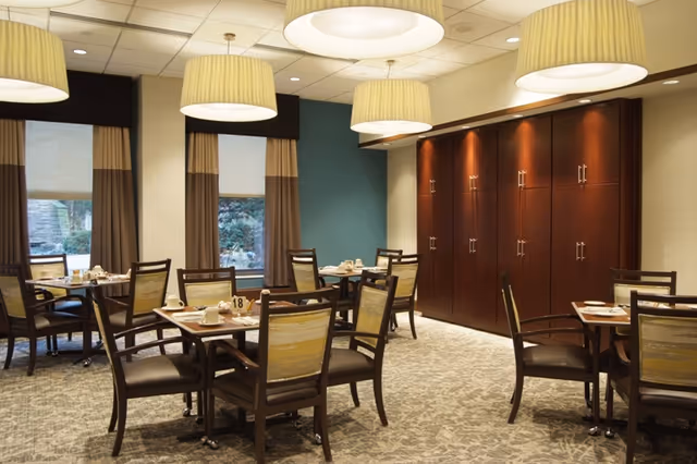 A dining room with several wooden tables and chairs arranged neatly. Each table is set with cups, plates, and utensils. The room has large windows with curtains, a patterned carpet, and modern ceiling lights. There is a wall with wooden cabinets in the background.