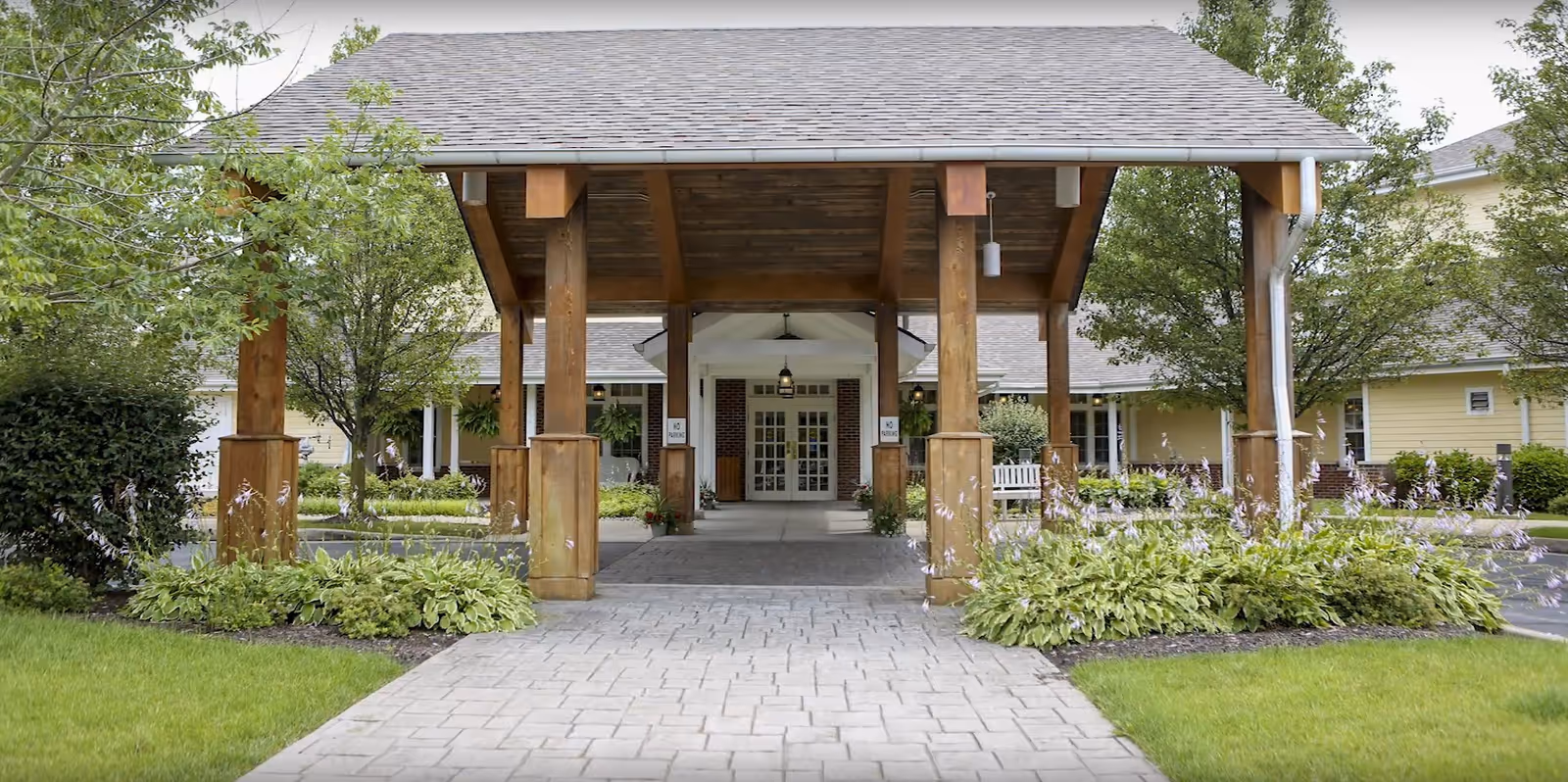 Entrance to River Terrace Retirement Community featuring a covered wooden porte-cochere with a paved walkway leading to double glass doors. The area is surrounded by green shrubs, trees, and well-maintained landscaping.