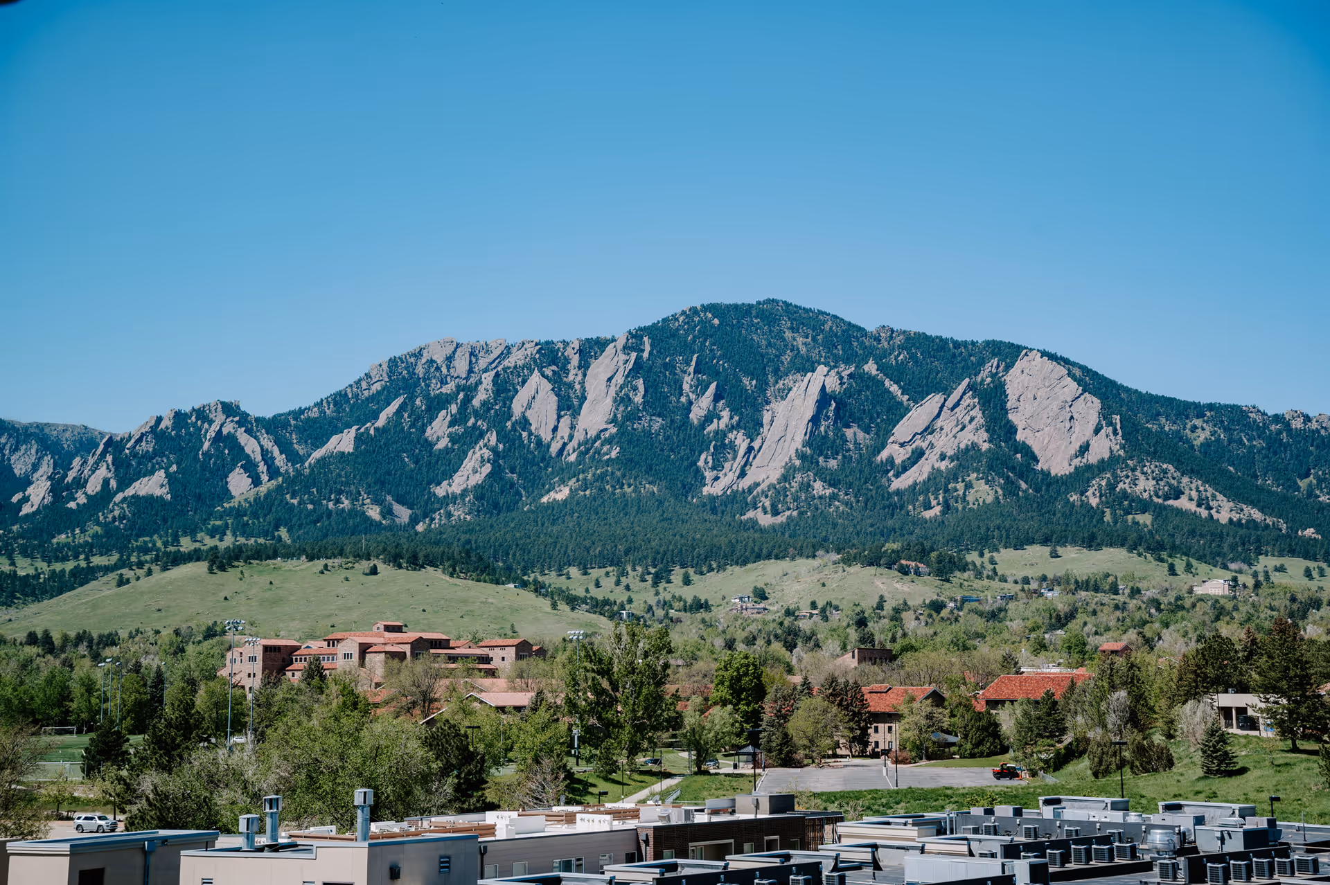 A scenic view of a mountain range with rocky formations and green forested areas under a clear blue sky. In the foreground, there are buildings with flat roofs and some trees scattered around.
