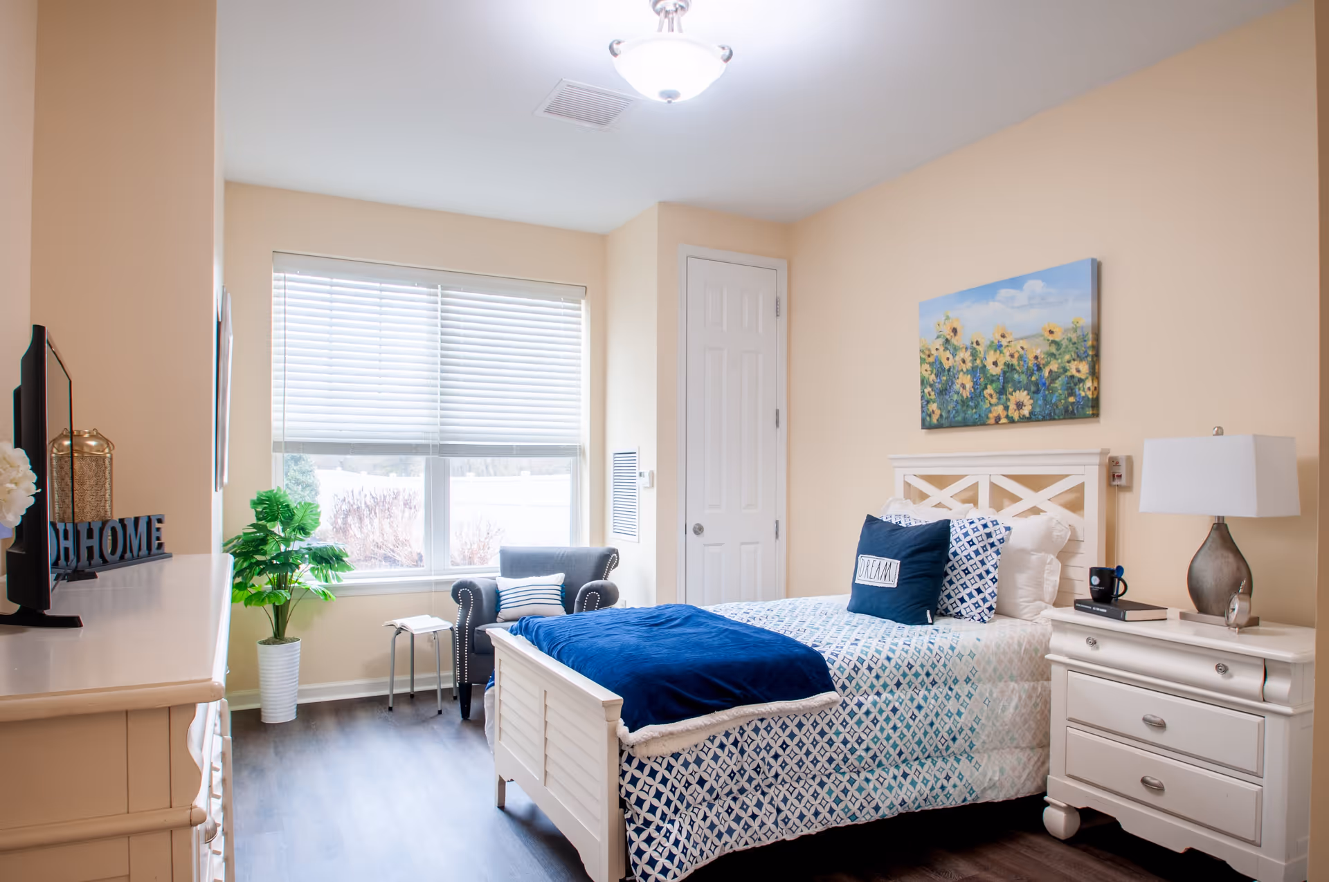 A bright and cozy bedroom with a single white bed featuring blue and white patterned bedding and a navy blue throw blanket. There is a white nightstand with a lamp, a book, and a coffee mug. A painting of sunflowers hangs above the bed. A gray armchair with a striped pillow sits near a window with white blinds. A white dresser with a TV and decorative items is on the left side of the room. The walls are painted beige and the floor is dark wood.