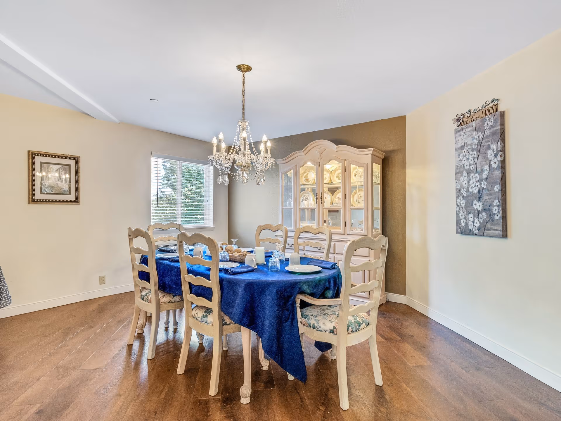 A dining room with a wooden floor, a rectangular table covered with a blue tablecloth, six wooden chairs with floral cushions, a chandelier hanging above the table, a window with blinds, a framed picture on one wall, a floral wall art on another wall, and a china cabinet displaying plates.