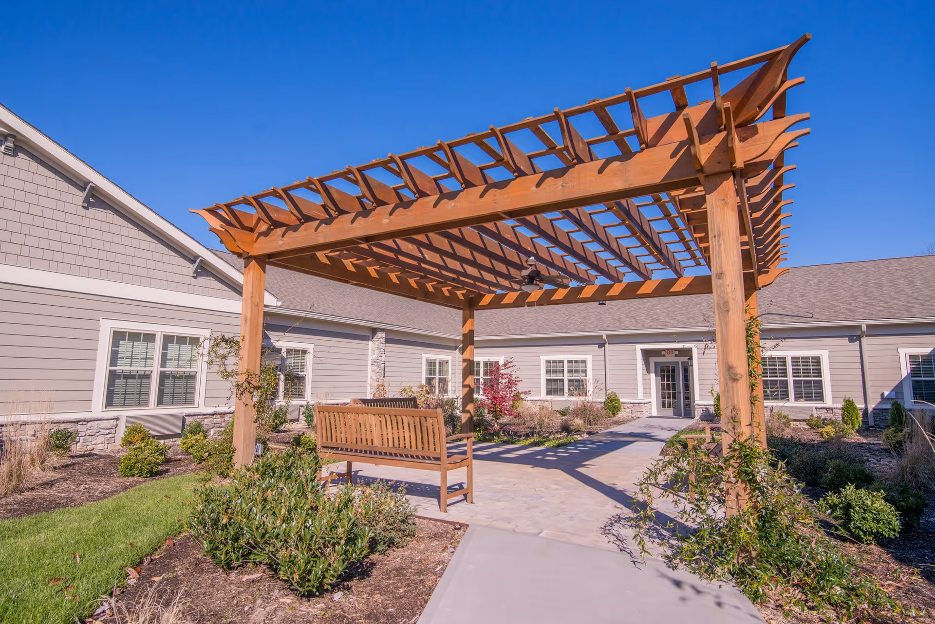 Outdoor courtyard area at Morning Pointe of Chattanooga at Shallowford featuring a wooden pergola with a bench underneath, surrounded by landscaped bushes and plants, with the facility building in the background under a clear blue sky.