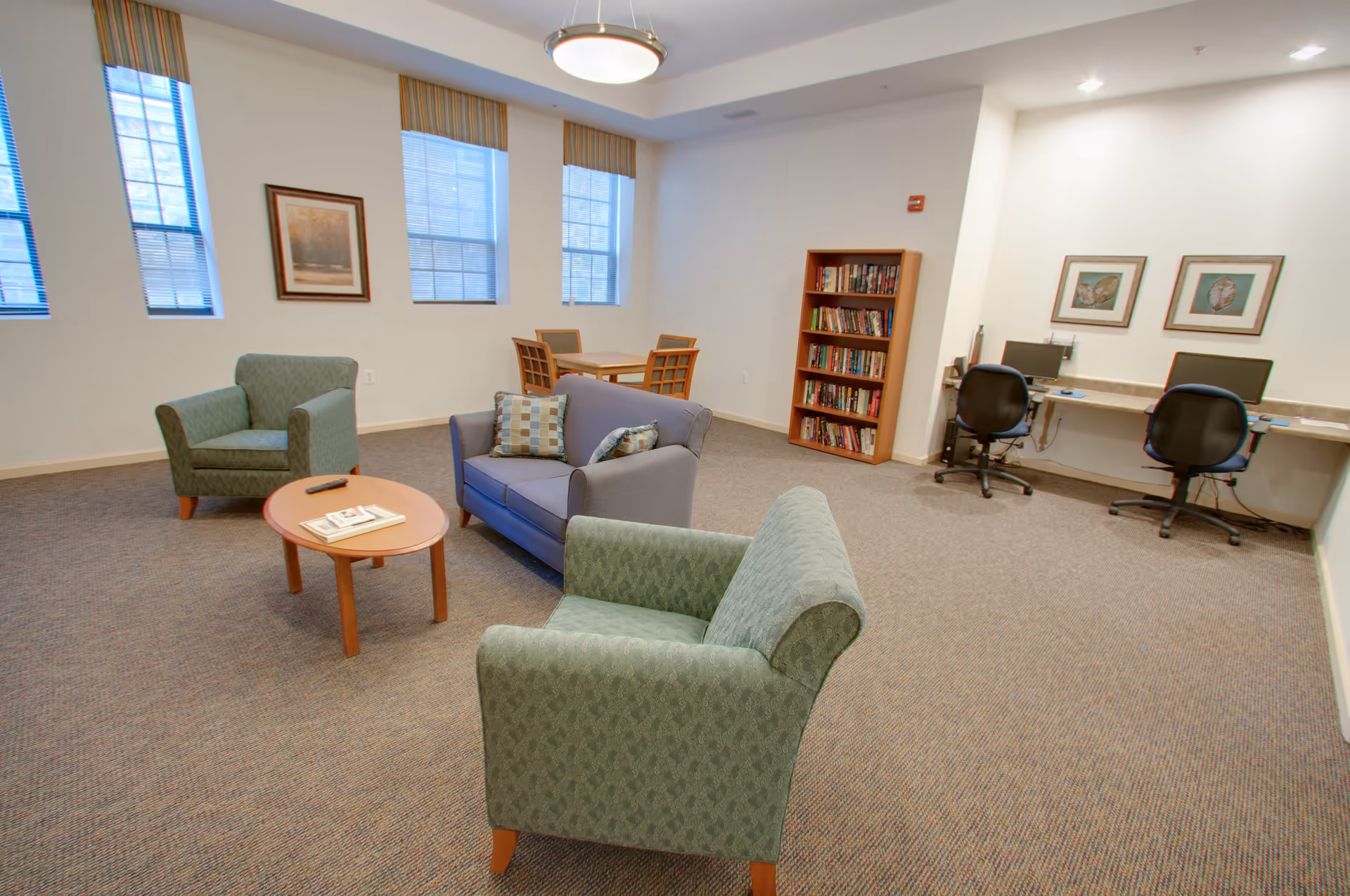 A common area in a senior living facility with two green armchairs, a blue loveseat with patterned pillows, a wooden coffee table with magazines and a remote, a small dining table with four chairs, a bookshelf filled with books, and two computer workstations with black office chairs. The room has large windows with blinds and framed artwork on the walls.