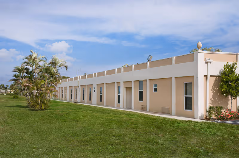 Long single-story beige building with windows and doors, a green lawn and palm trees under a blue sky.