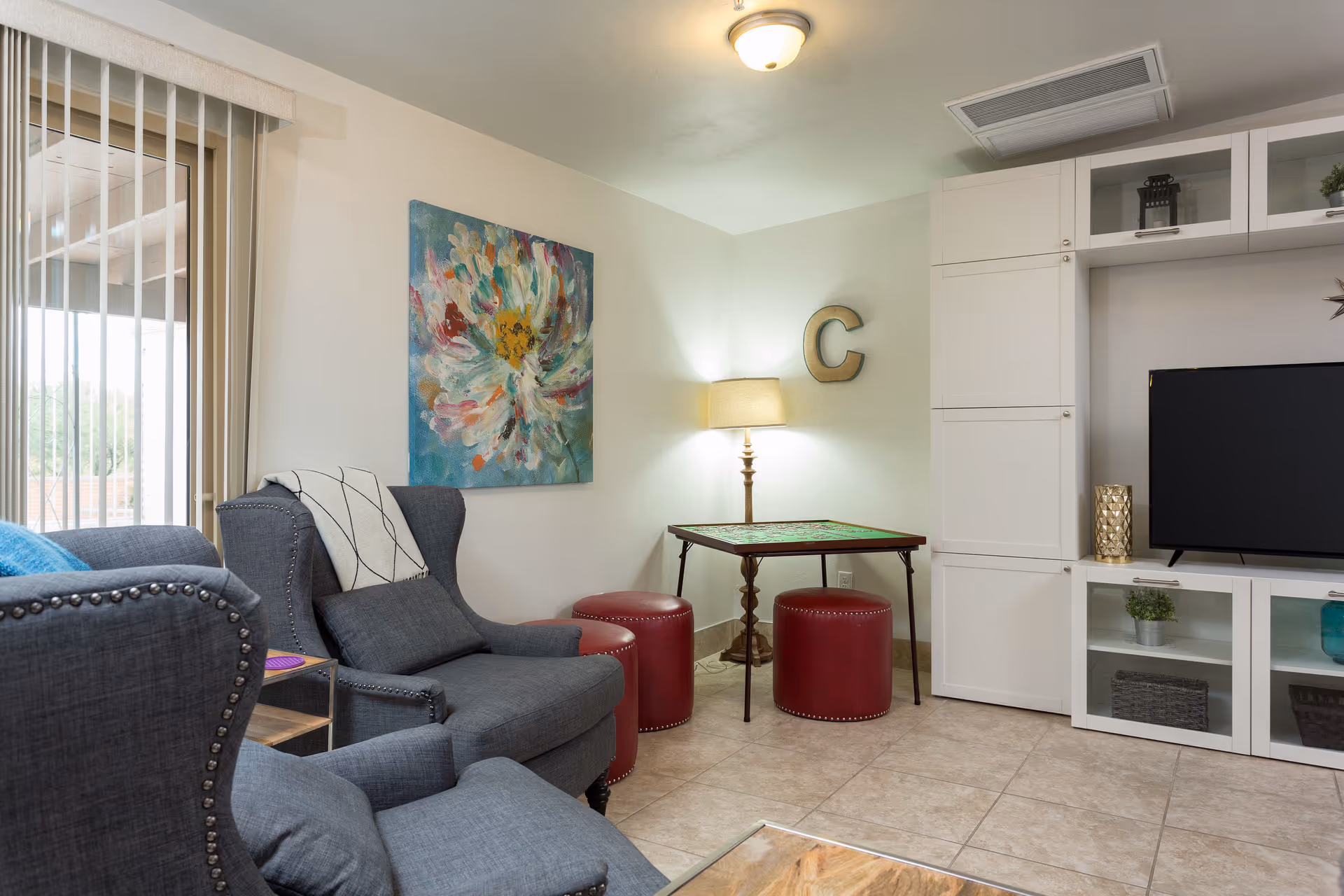 A cozy living room with two gray armchairs, a small wooden side table, a colorful floral painting on the wall, a lamp next to a small table with a puzzle on it, three red round stools, and a white entertainment unit with a flat-screen TV and decorative items.
