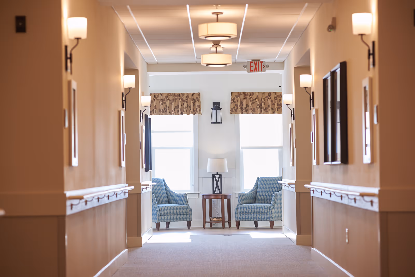 A well-lit hallway leading to a seating area with two patterned armchairs and a side table beneath windows.