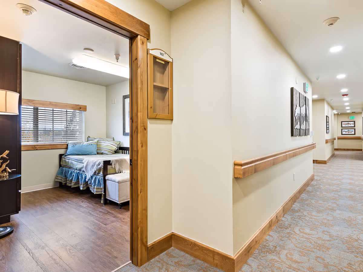 View of a hallway in a senior living facility with beige walls, wooden trim, and carpeted floor. On the left, there is an open doorway leading to a bedroom with a bed, pillows, a window with blinds, and a lamp on a side table.