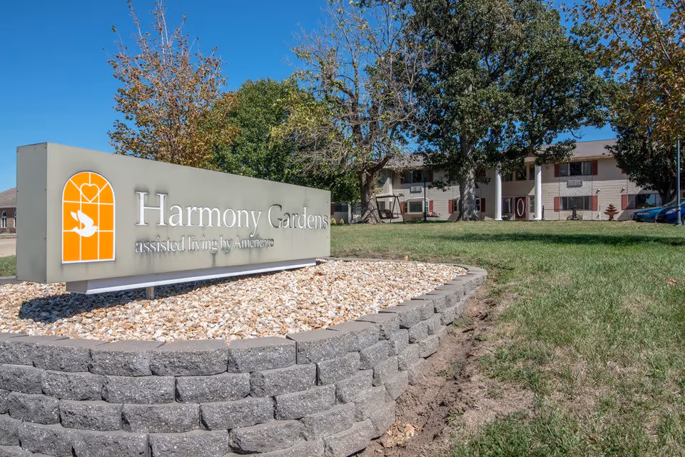 Outdoor view of Harmony Gardens assisted living facility sign with a building and trees in the background under a clear blue sky.