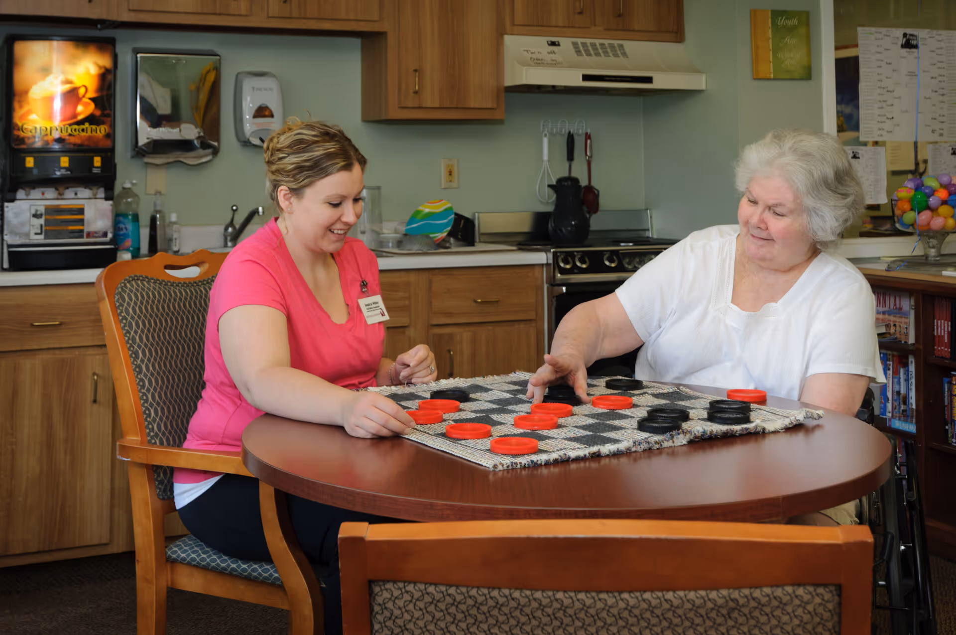 Two women sit at a round table in a common-area kitchen playing checkers.