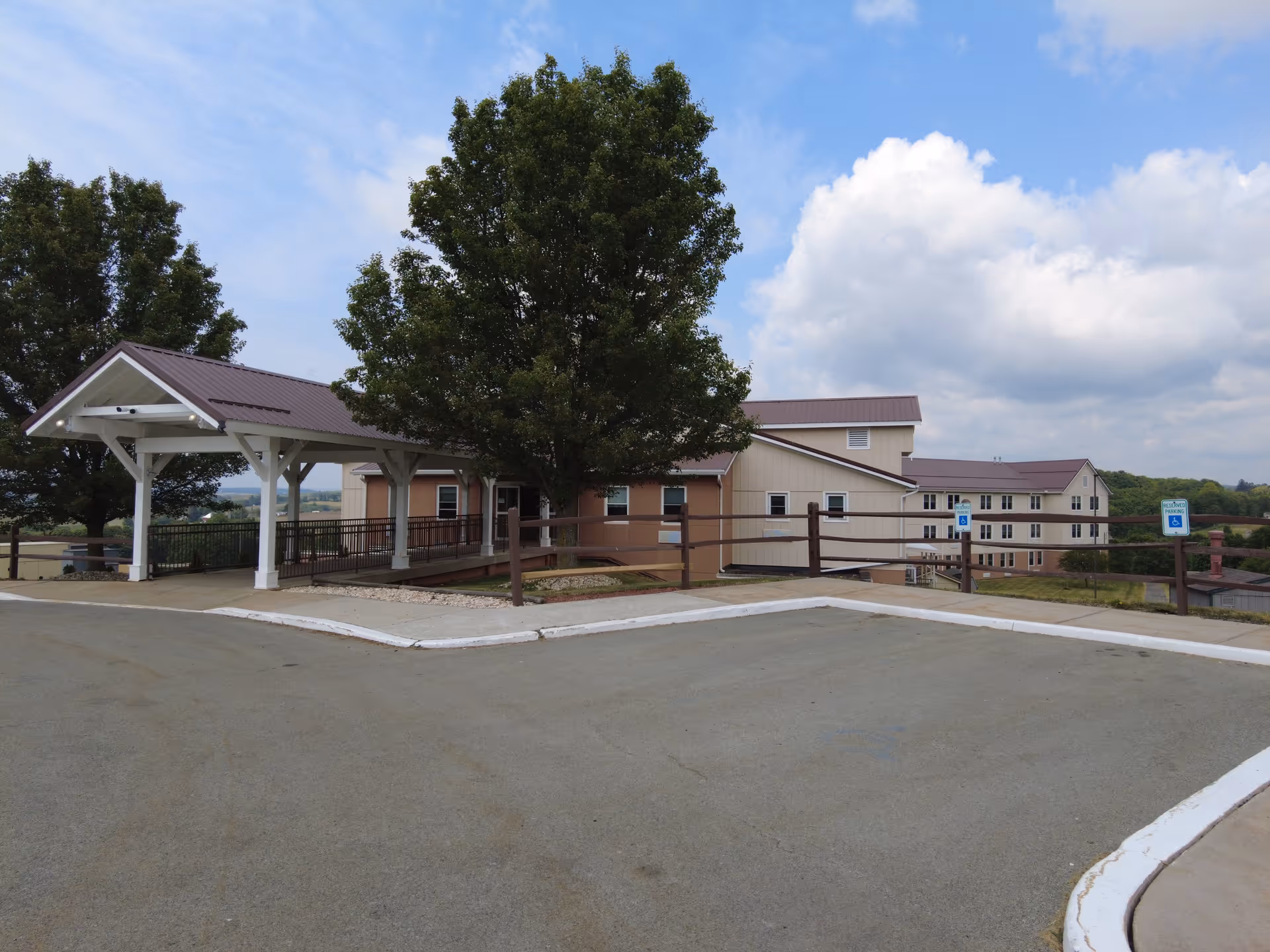 View of the exterior of Meadow View Nursing Center showing a multi-story building with beige and brown siding, a covered entrance with white pillars, a tree in front, and a parking area with designated handicapped parking spaces under a partly cloudy sky.