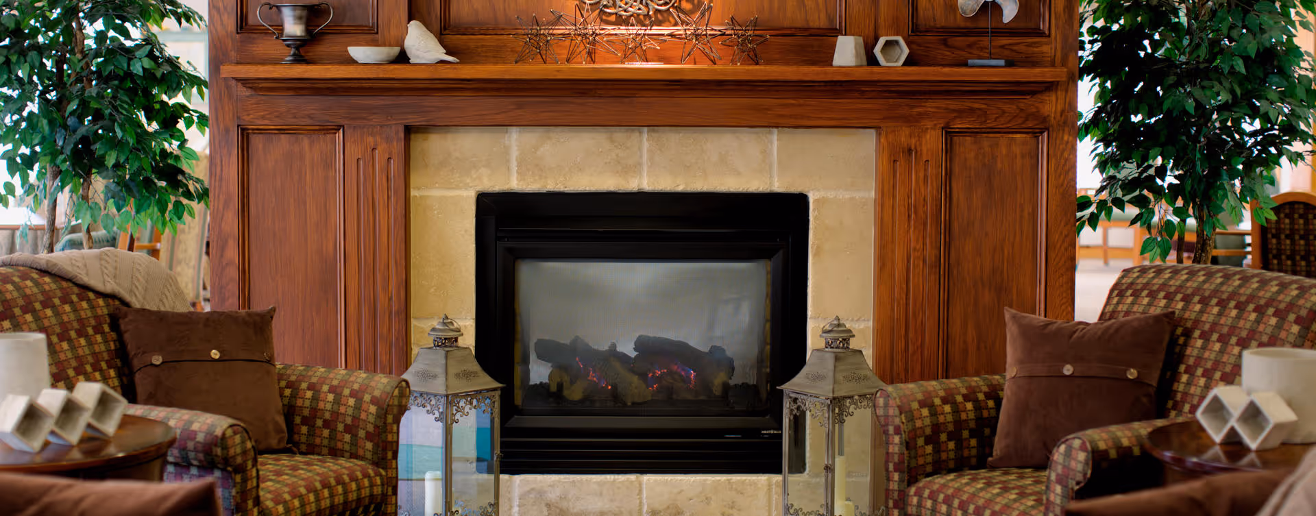 A cozy living room area featuring a central fireplace with a wooden mantel, flanked by two patterned armchairs with brown cushions. Two decorative lanterns are placed on the floor in front of the fireplace, and there are green plants on either side of the seating area. The background shows a glimpse of a dining area with chairs and tables.