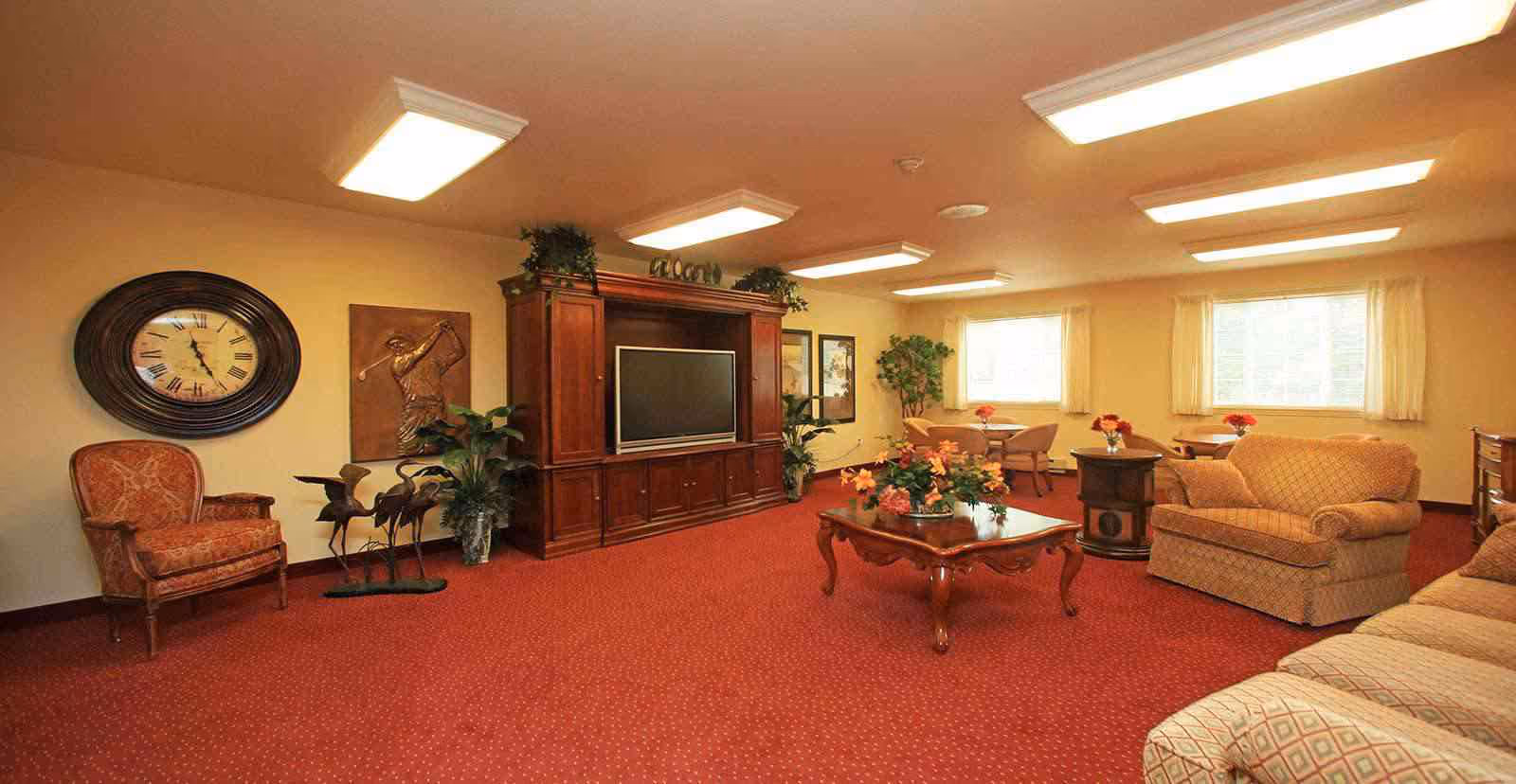 Communal living room with patterned carpet, upholstered sofas and chairs, a wooden entertainment center with TV, and a central coffee table with flowers.
