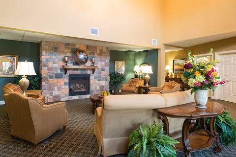 A cozy living room area in a senior living facility featuring a stone fireplace with a decorative globe on the mantel, surrounded by comfortable upholstered chairs and sofas. The room is warmly lit with table lamps and decorated with framed artwork, potted plants, and a large floral arrangement on a wooden side table.