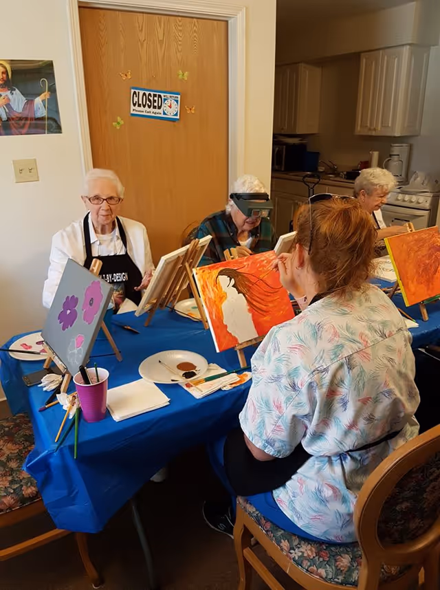 Four elderly women seated around a table covered with a blue tablecloth, engaged in painting on canvases set on easels. The room appears to be a kitchen or dining area with cabinets, a stove, and a refrigerator in the background. One woman is painting a figure with flowing hair on an orange background, while another canvas shows purple flowers. A closed wooden door with a 'CLOSED Please Call Again' sign is visible behind them.