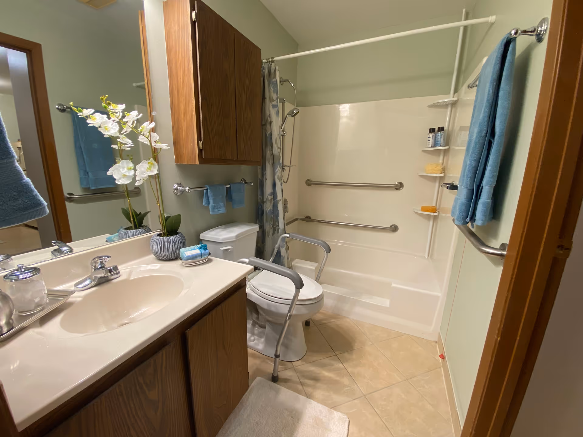 A bathroom with a sink and countertop on the left, a toilet with safety rails in the center, and a bathtub with grab bars and a shower curtain on the right. There are blue towels hanging on the walls and a vase with white flowers on the countertop.