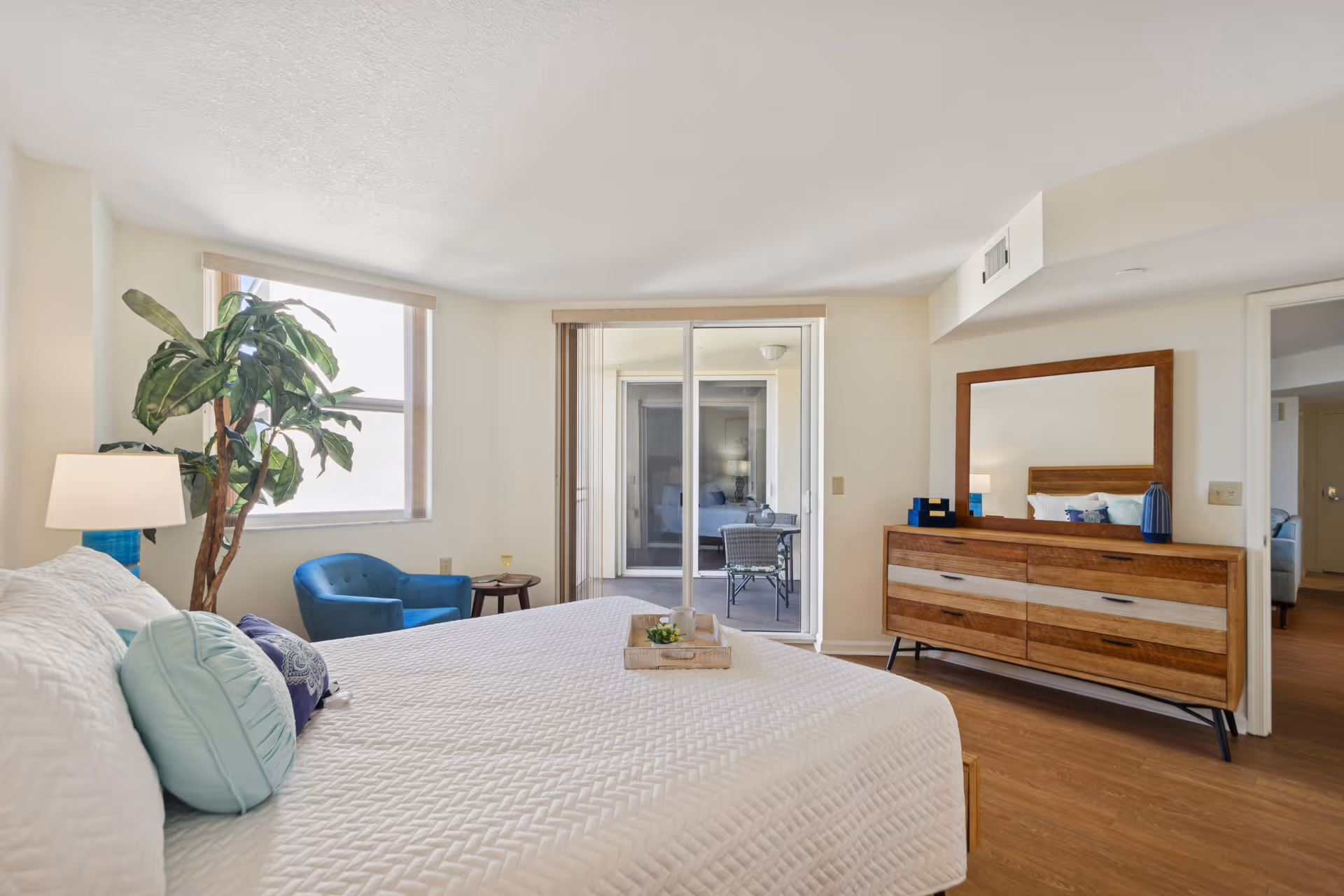 Bright and spacious bedroom with a large bed covered in white bedding and multiple pillows. A blue armchair and small round table are placed near a window with natural light. A tall green plant is beside the bed. A wooden dresser with a large mirror stands against the wall, and sliding glass doors lead to a small outdoor seating area with a table and chairs.