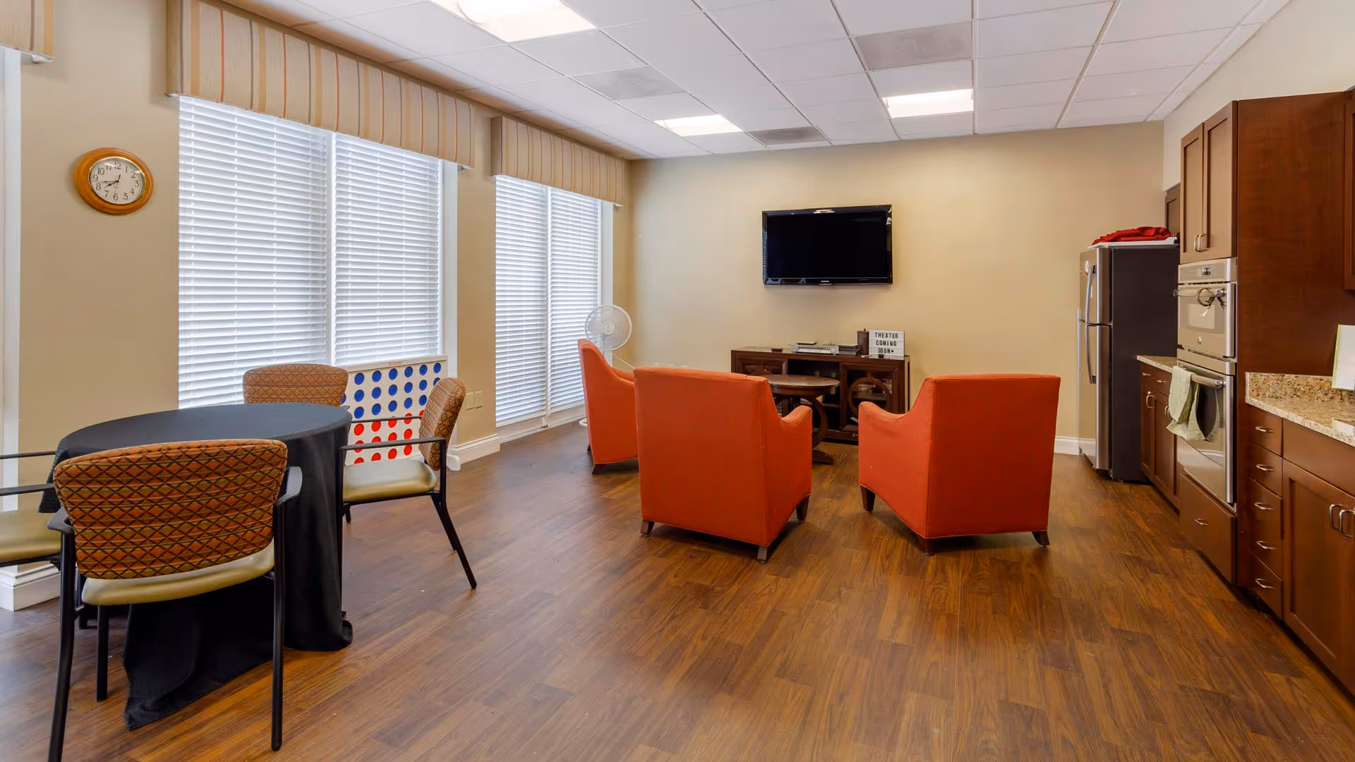 A communal room with a small kitchen area featuring wooden cabinets, a refrigerator, and an oven. There are three orange armchairs arranged facing a wall-mounted flat-screen TV. To the left, there is a round table with four chairs and a large Connect Four game near the windows with blinds. The room has wood flooring and a clock on the wall.