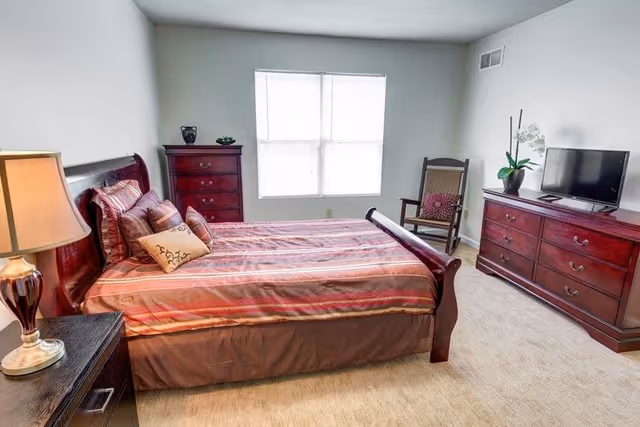 A bedroom with a large wooden bed featuring a striped bedspread and multiple pillows. There is a wooden nightstand with a lamp on the left side of the bed, a tall wooden dresser against the far wall, a wooden rocking chair with a cushion near the window, and a long wooden dresser with a flat-screen TV and a potted plant on top. The room has beige carpet and a window with blinds letting in natural light.