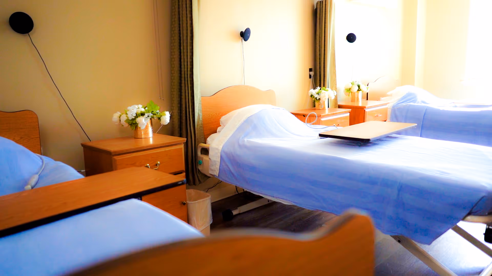 A room with three hospital-style beds covered in light blue sheets, each with a wooden bedside table holding a small vase of white flowers. The room has beige walls, green curtains, and wall-mounted black round lights above each bed.