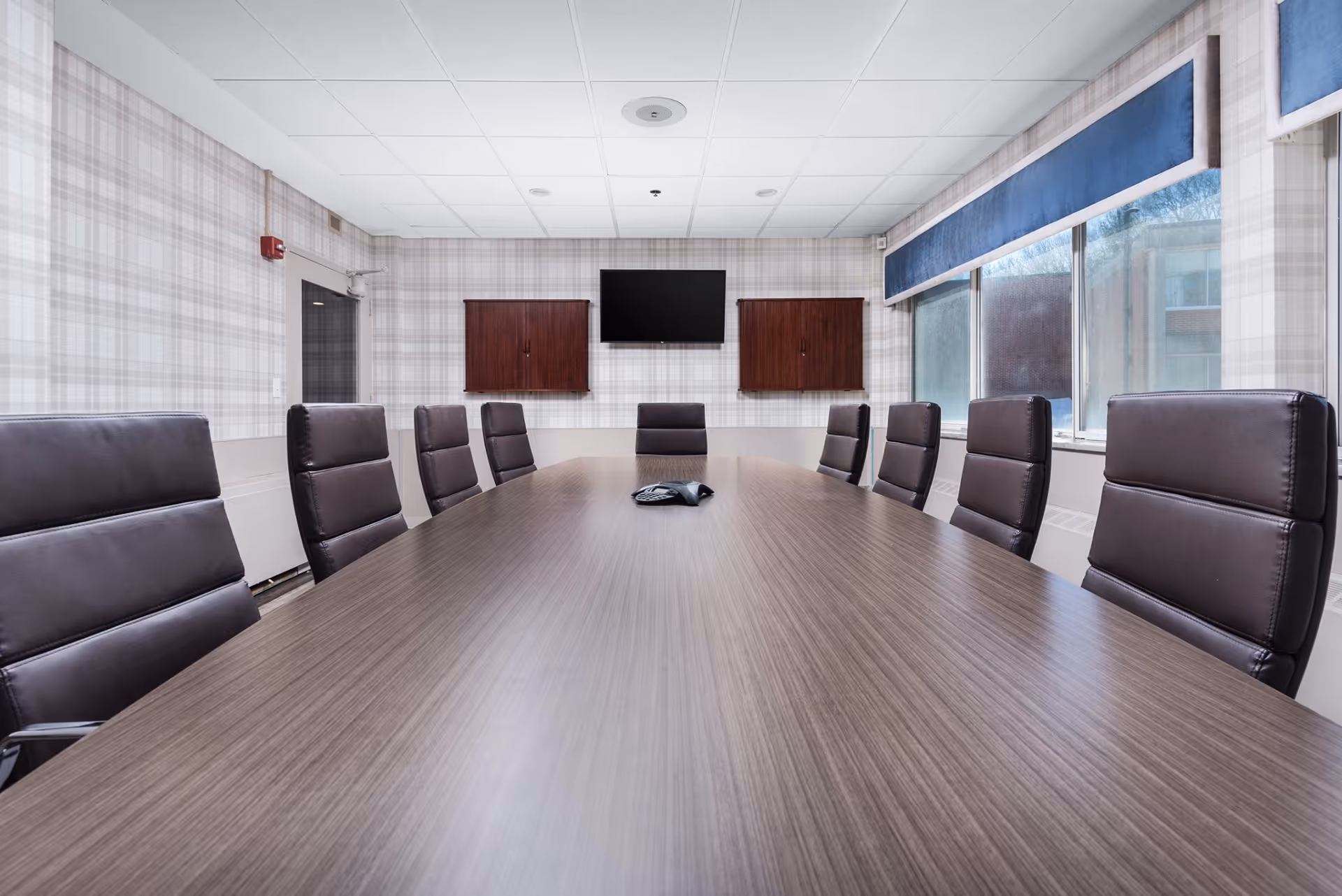 A modern conference room with a long wooden table surrounded by black leather chairs. The room has plaid wallpaper, a wall-mounted flat screen TV, two wooden cabinets, and large windows with blue valances letting in natural light.