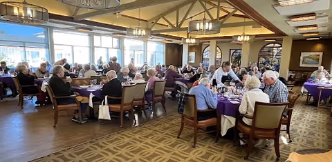 A spacious dining room filled with elderly residents seated at multiple round tables covered with purple tablecloths. A waiter is serving food to one of the tables. Large windows on one side let in natural light, and the room features warm lighting fixtures and carpeted flooring.