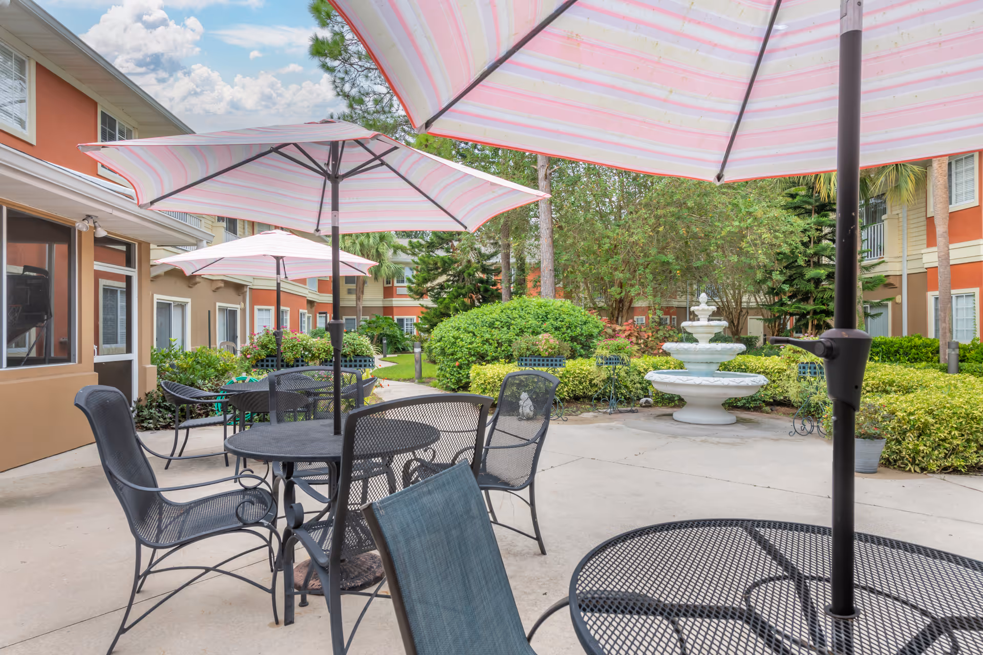 Courtyard patio with metal tables and chairs shaded by pink-striped umbrellas and a central fountain in front of apartment-style buildings.