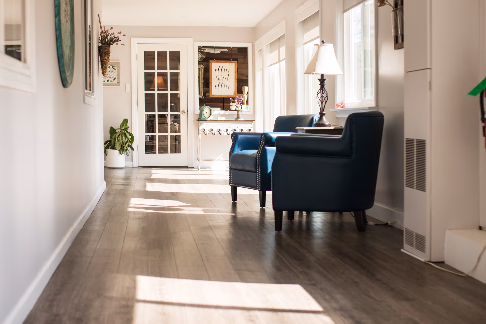 A bright hallway with wooden flooring featuring two blue armchairs and a side table with a lamp near windows on the right. At the end of the hallway, there is a white door with glass panels and a small table with decorative items and a framed sign that reads 'office sweet office'. A potted plant is placed on the left side near the door.