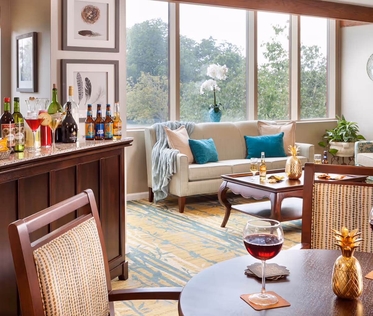 Sunny common lounge with large windows, a sofa with teal pillows, a coffee table, a dining table with a glass of red wine, and a small bar stocked with bottles.