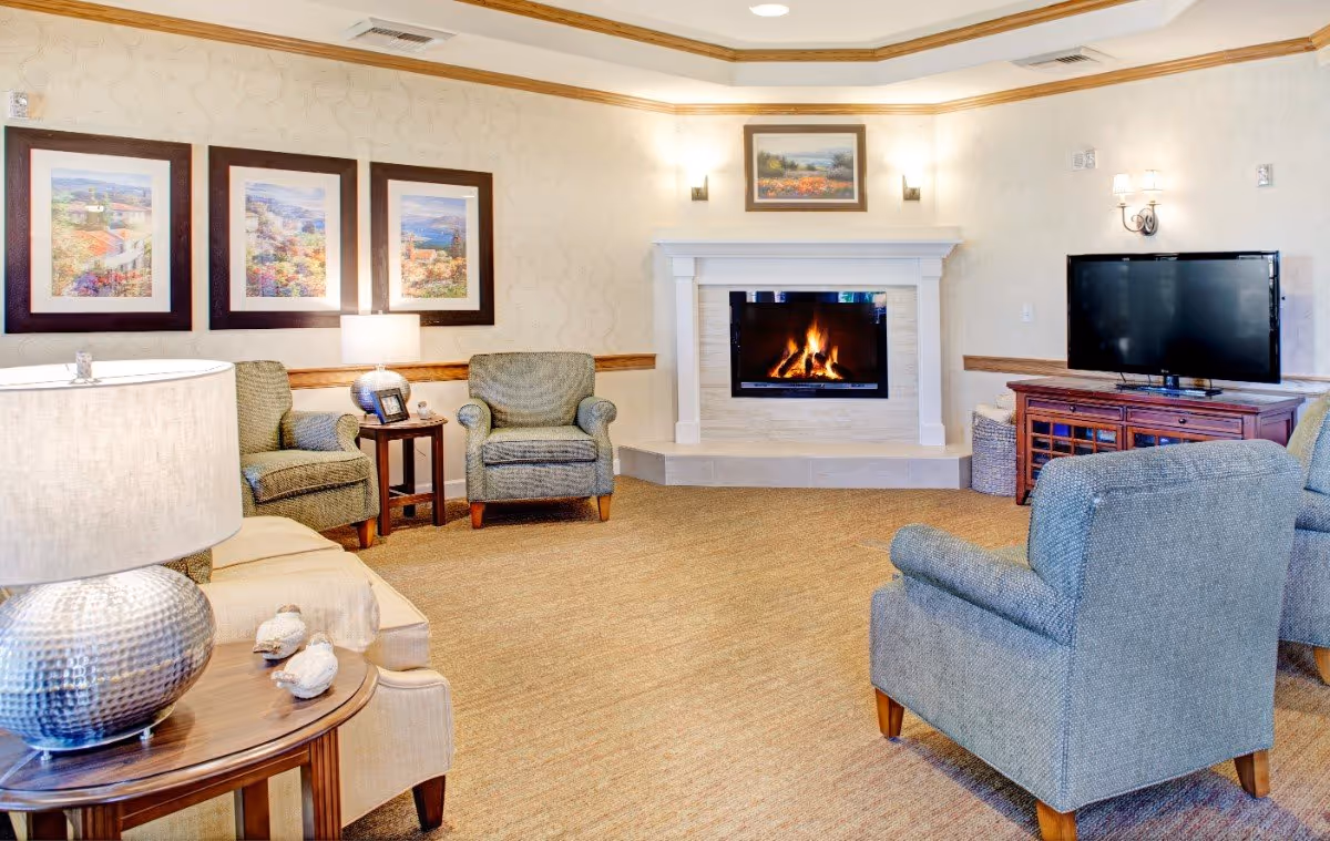 A cozy living room with a lit fireplace centered on the far wall, surrounded by four upholstered armchairs and a beige sofa. The room features a flat-screen TV on a wooden cabinet to the right, a round wooden side table with a silver lamp and decorative birds on the left, and three framed landscape paintings on the wall above the chairs. The walls have a light patterned wallpaper with wooden trim, and the floor is carpeted.
