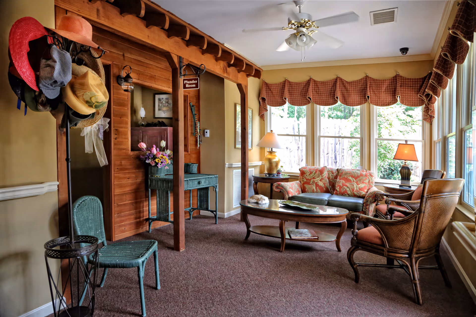 A cozy living room area with a green wicker chair and a hat rack holding various hats on the left. The room features a wooden wall with a mirror and a small table with flowers. On the right, there is a seating area with a floral-patterned sofa, two armchairs, a wooden coffee table, and two table lamps. Large windows with red valances allow natural light to fill the room.