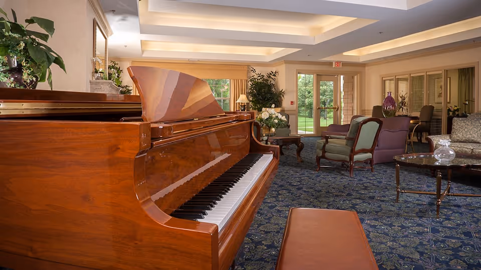 Interior view of a senior living facility lounge area featuring a polished wooden grand piano in the foreground, upholstered chairs and sofas arranged around glass-top coffee tables, decorative plants, and large windows with a view of greenery outside.