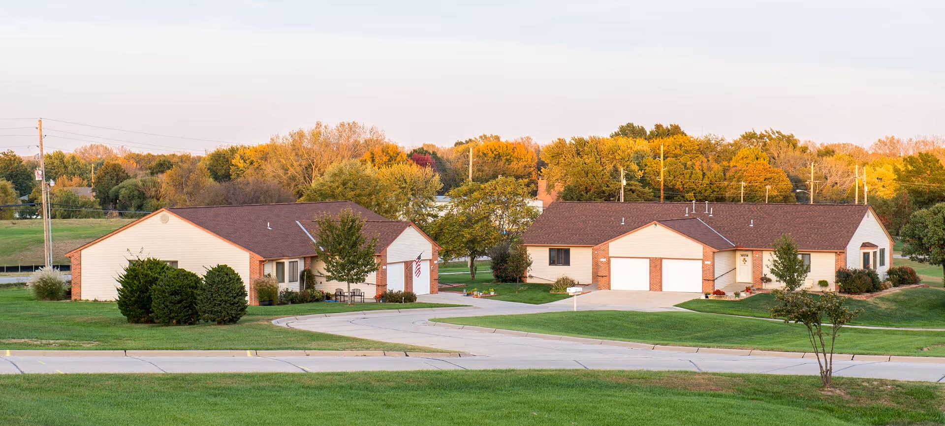 Two single-story residential buildings with beige siding and brown roofs set in a green grassy area with trees showing autumn colors in the background under a clear sky.