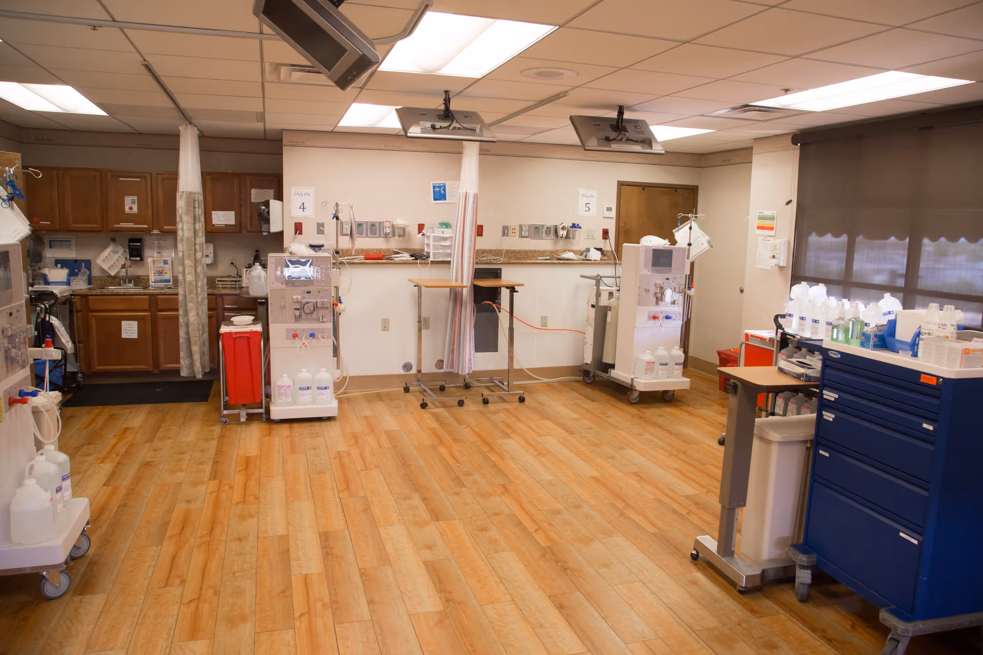 A clean medical treatment room with wooden flooring, medical equipment on carts, cabinets along the back wall, and privacy curtains. The room is well-lit with ceiling lights and has monitors mounted on the ceiling.
