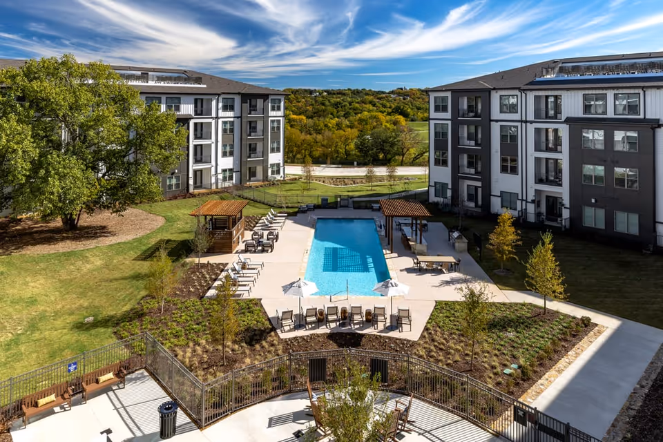 Outdoor view of a senior living facility courtyard featuring a rectangular swimming pool surrounded by lounge chairs, umbrellas, and two shaded pergolas. The courtyard is bordered by two multi-story residential buildings with large windows. Trees and landscaped greenery surround the pool area under a partly cloudy blue sky.