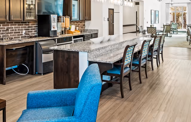 Communal dining and kitchenette area with a long granite island, bar stools, beverage dispensers, and blue upholstered seating.