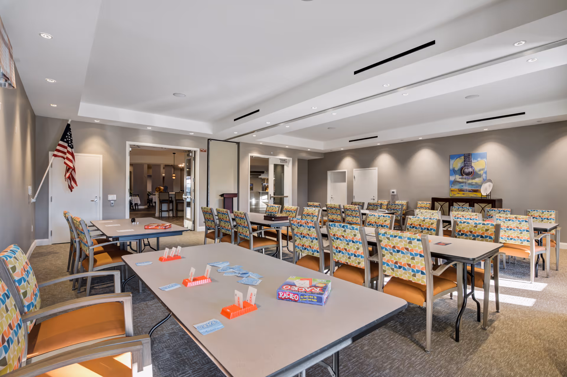 A bright multipurpose dining/activity room with rows of tables, colorful patterned chairs, and game supplies on the tables.