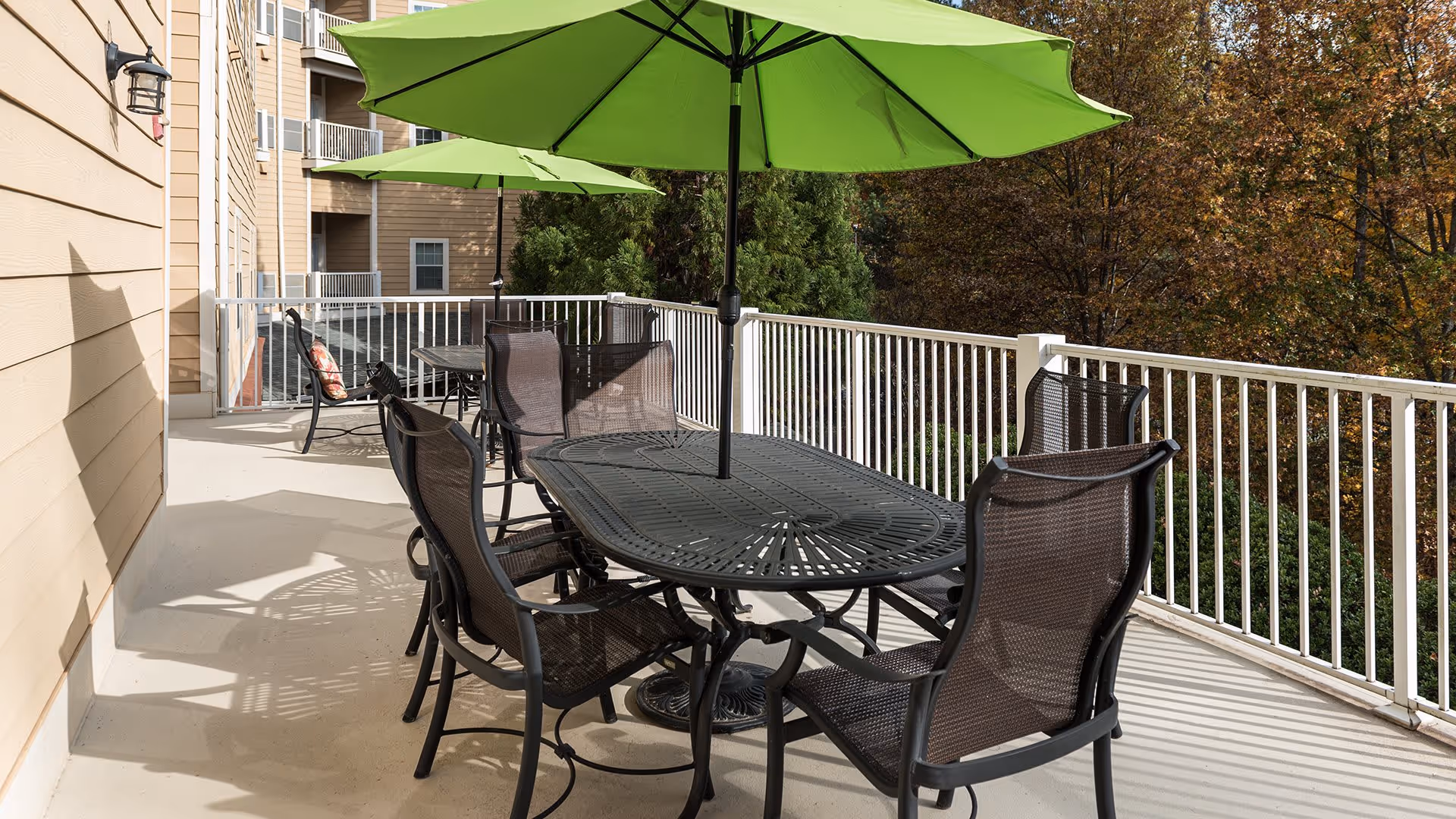 Outdoor patio area with black metal tables and chairs, each table shaded by a large green umbrella. The patio is surrounded by a white railing and overlooks trees with autumn foliage.