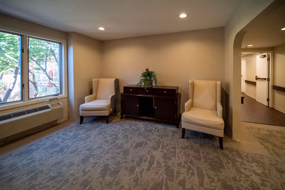 A small sitting area in a senior living facility with two beige upholstered armchairs placed on either side of a dark wooden cabinet with a potted plant on top. The room has a large window showing green trees outside, beige walls, recessed ceiling lights, and a patterned carpet. A hallway with wooden flooring and white doors is visible through an archway on the right.