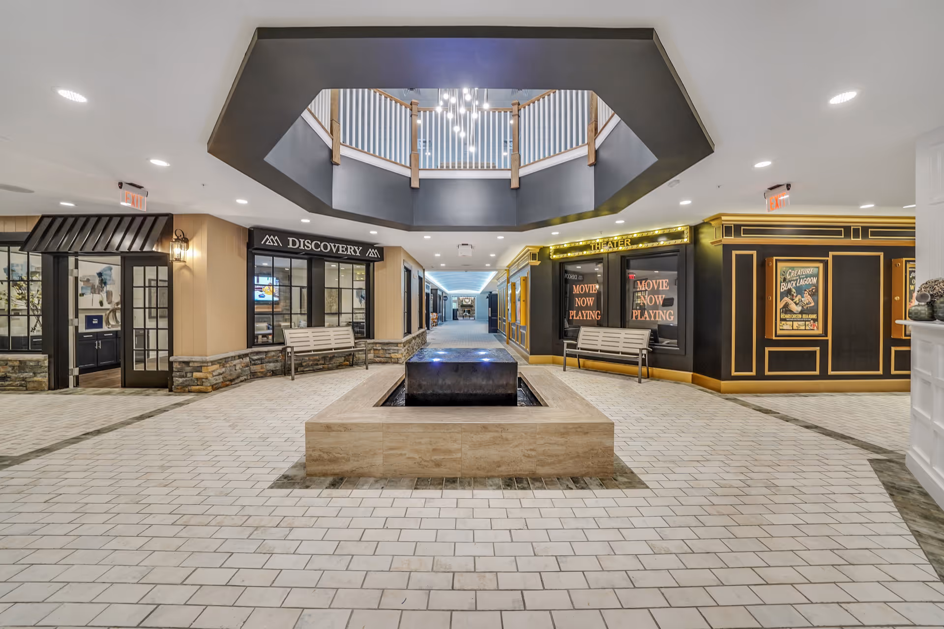 Interior view of a senior living facility hallway with a central water feature, benches, and storefront-style entrances labeled 'DISCOVERY' and 'THEATER' with movie posters. The ceiling features a modern chandelier and an open balcony above.