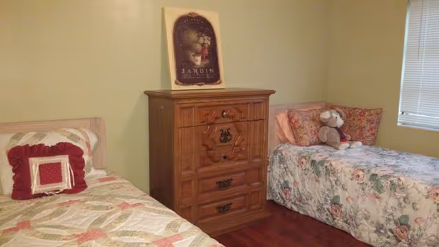 Two beds with floral quilts flank a wooden dresser; a teddy bear sits on right bed.