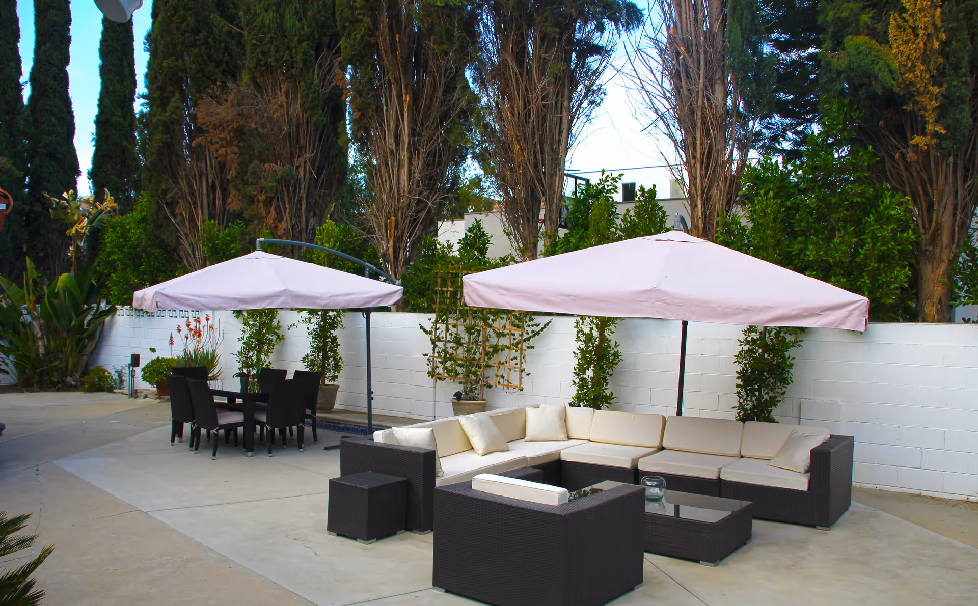 Outdoor patio area with two large pink umbrellas providing shade over a black wicker sectional sofa with cream cushions and a glass coffee table, and a black dining table with six chairs. The area is surrounded by tall trees and a white brick wall with potted plants.
