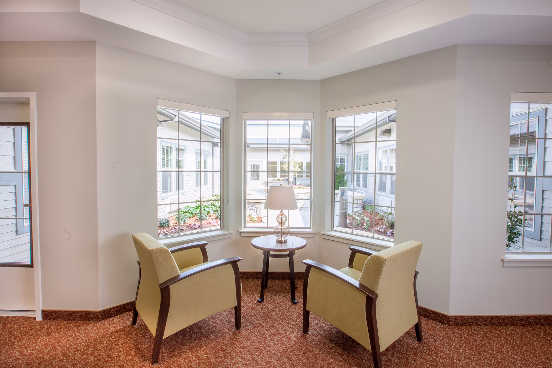 A cozy seating area with two beige armchairs facing each other and a small round wooden table with a lamp between them, positioned in front of three large windows overlooking a courtyard with plants and pathways.